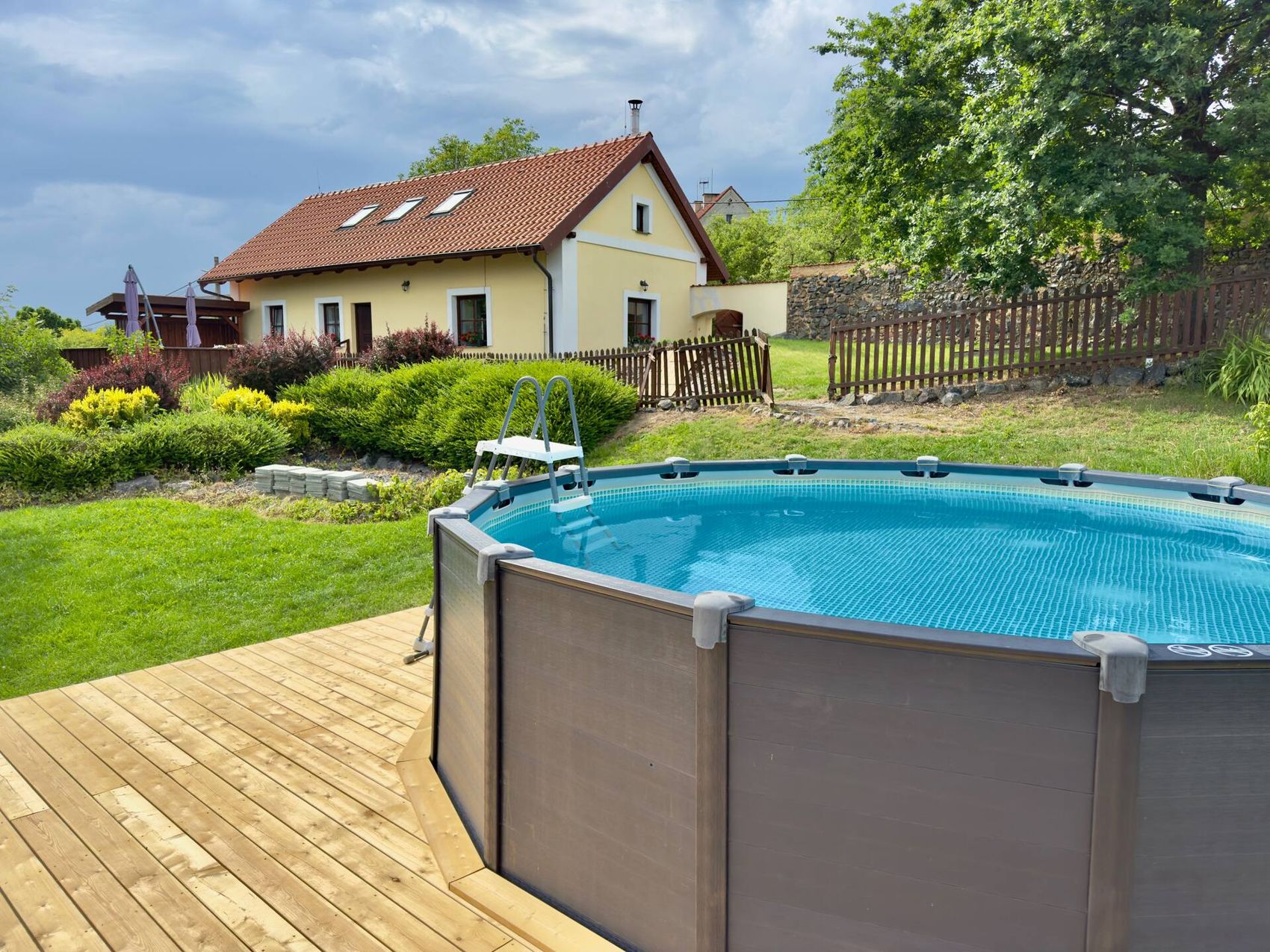 Nadzemní bazén na dřevěné terase, za ním žlutá chalupa a zahrada